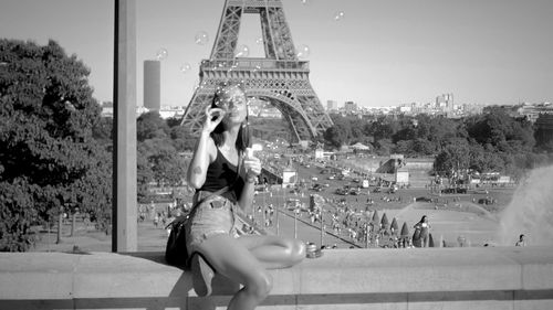 Woman blowing bubbles while sitting on retaining wall against eiffel tower