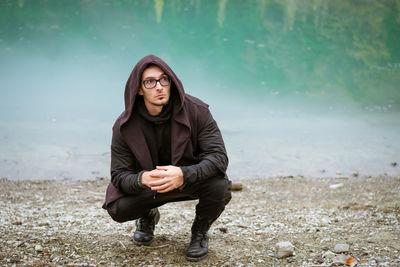 Bearded man in casual clothes with hood and glasses stands on shore