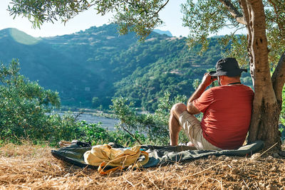 Rear view of tourist man resting in olive grove, looking through binoculars at the mountains view. 