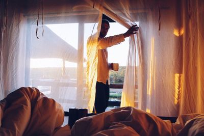 Man holding coffee cup looking through curtain at home