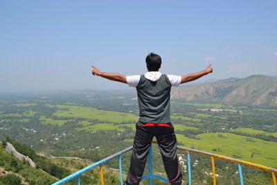 Rear view of man with arms outstretched standing on railing against mountains and sky