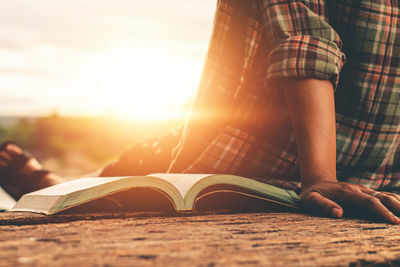 Midsection of person reading book at beach against sky during sunset