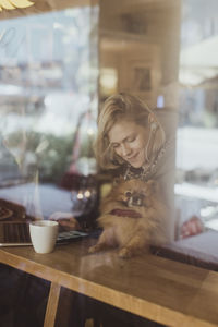 Smiling blond woman sitting with pomeranian at cafe seen through window