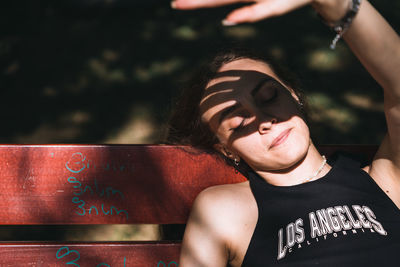 Low angle view of young woman sitting on bench and shadowing the sun