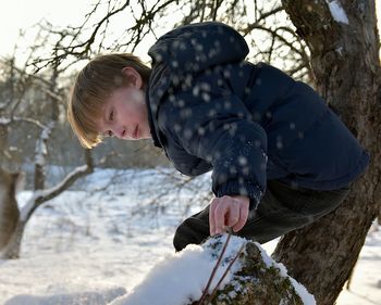 Side view of boy bending by bare tree on snow covered field