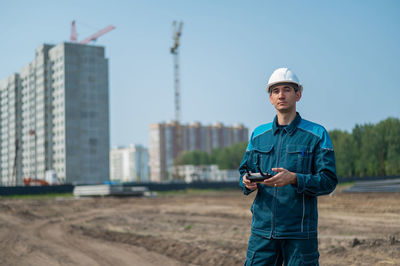 A man in a helmet and overalls controls a drone at a construction site. the builder carries out