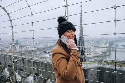 Portrait of young woman using mobile phone standing by railing in city during winter
