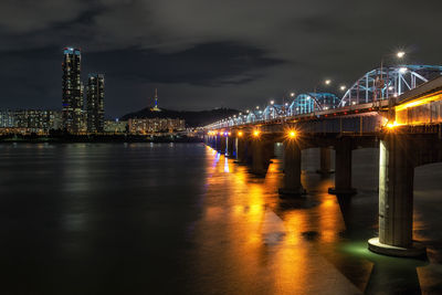 Illuminated bridge over river against buildings at night