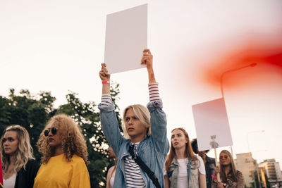 Young men and women protesting with posters while marching in city against sky