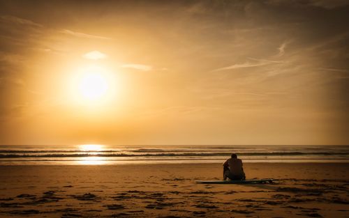 Man sitting on beach against sky during sunset
