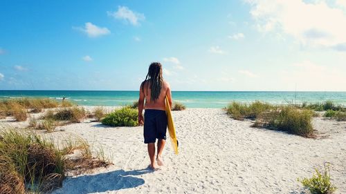 Rear view of shirtless man holding surfboard walking on shore at beach against sky