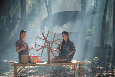 Women using spinning wheel while sitting on wooden bench outdoors