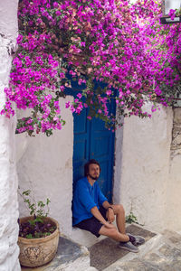 Man in a blue shirt with a beard sitting on  steps next to the blue door and pink flowers on a bush