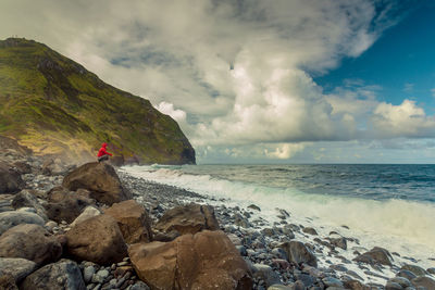 Side view of woman crouching on rock against cloudy sky