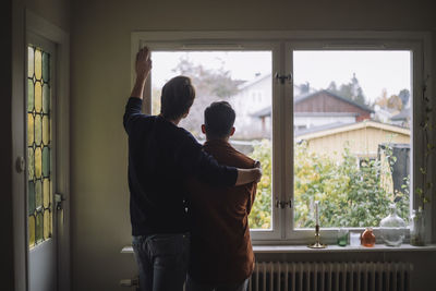 Rear view of man with arm around gay couple looking through window at home