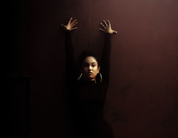 Portrait of young woman standing with arms raised against wall