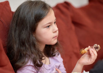 Expressive young girl is eating chicken and vegetables for dinner on the couch while watching tv