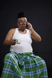 Mature man talking on phone while holding coffee cup against black background