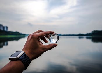 Cropped hand holding heart shape diamond by lake against cloudy sky