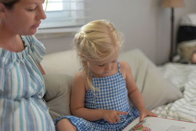 Mother looking at cute daughter reading book while sitting on bed