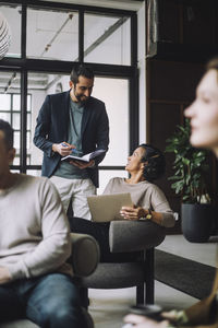 Smiling businessman holding diary while discussing with female colleague sitting on chair in creative office