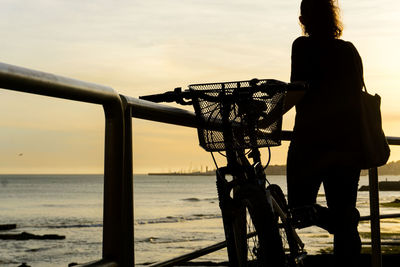 Man standing on bicycle by sea against sky during sunset