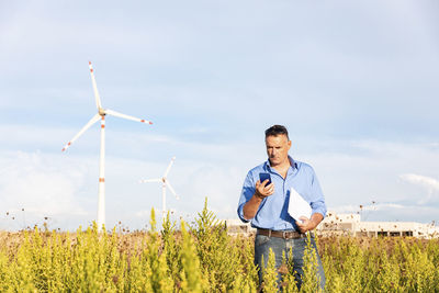 Mature businessman holding smart phone and document while standing against wind turbines on field