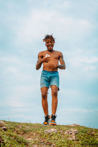 Full length of shirtless man holding a mobile phone standing against sky