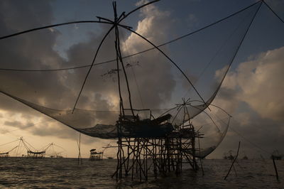 Silhouette chinese fishing nets in sea against cloudy sky