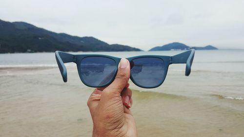 Cropped hand holding sunglasses by sea against sky