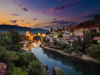 Bridge over river by buildings against sky during sunset