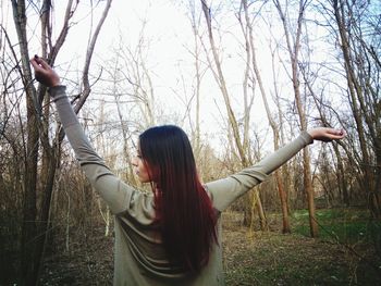 Rear view of young woman standing with arms outstretched against bare trees