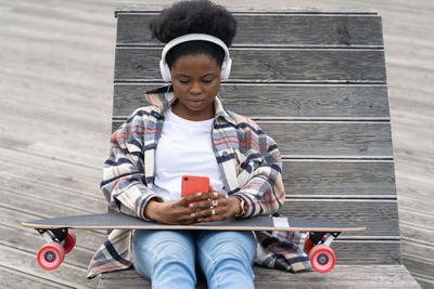 Serious african woman skater texting in smartphone with longboard on knees sit with skate on bench