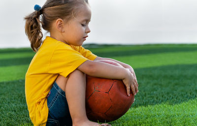 Cute little girl sitting with ball on green lawn and looking at it