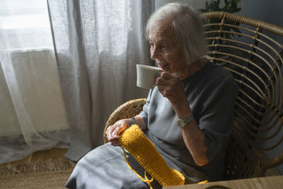 Senior woman having tea holding knitting wool on chair at home