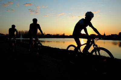 Silhouette people riding bicycle by lake during sunset