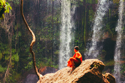 Rear view of man sitting by waterfall in forest