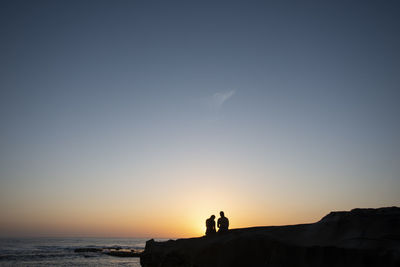 Silhouette people standing on sea shore against sky during sunset