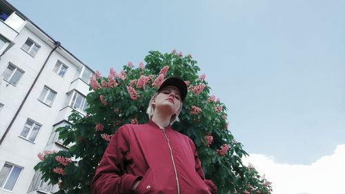 Low angle portrait of young woman standing by tree against sky in city