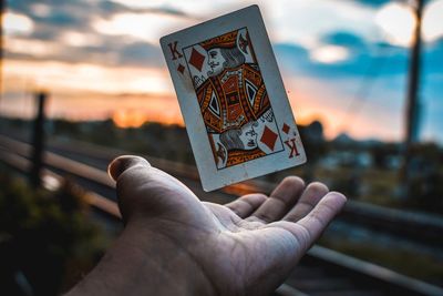 Close-up of hand catching king card against sky during sunset