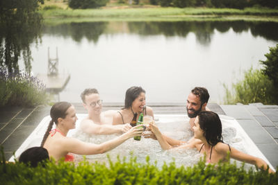 Cheerful male and female friends toasting drinks in hot tub against lake during weekend getaway