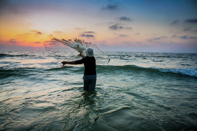 Rear view of man throwing fishing net in sea during sunset