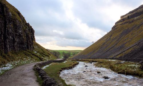 Empty dirt road by stream amidst mountain against cloudy sky