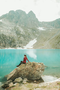 Man sitting on rock by lake against mountains