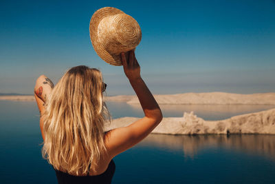 Rear view of woman holding hat while standing at beach against blue sky