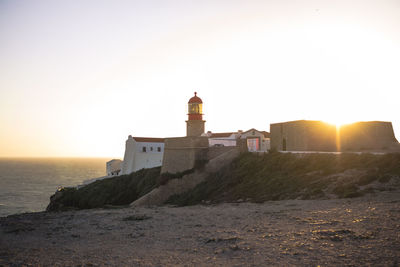 Lighthouse on cliff by sea against clear sky during sunset