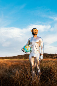 Anonymous guy in silver latex suit with geometric monkey mask looking away and holding globe in nature