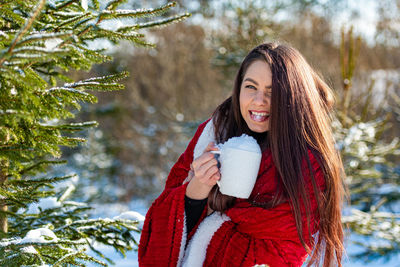 Teenage woman with a red blanket on her shoulder eating snow from a white cup in winter wood