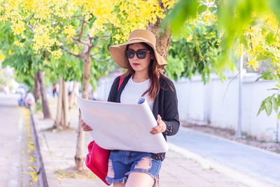 Portrait of young woman with backpack holding map while standing on road