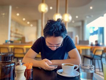Young asian man in eyeglasses using mobile phone while drinking coffee at table in cafe.